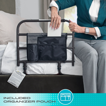A person sits on a bed, placing a book into the included black mesh organizer pouch attached to the metal Stander Wonder EZ Adjust Bed Rail - Extendable. The image features text that reads, Included Organizer Pouch.