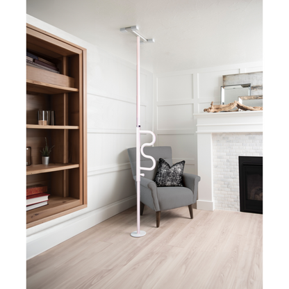 A Stander Wonder Pole - Fall Prevention Safety Pole For The Home extends from floor to ceiling beside a gray armchair in a bright living room with wooden floors, built-in shelves, and a white fireplace, providing convenient sit-to-stand support.
