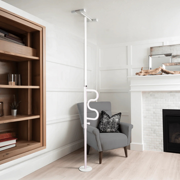 A Stander Wonder Pole HD - Bariatric Fall Prevention Safety Pole with a curved grab bar is installed beside a gray armchair in a modern living room with white walls, wooden shelving, and a fireplace for added support.