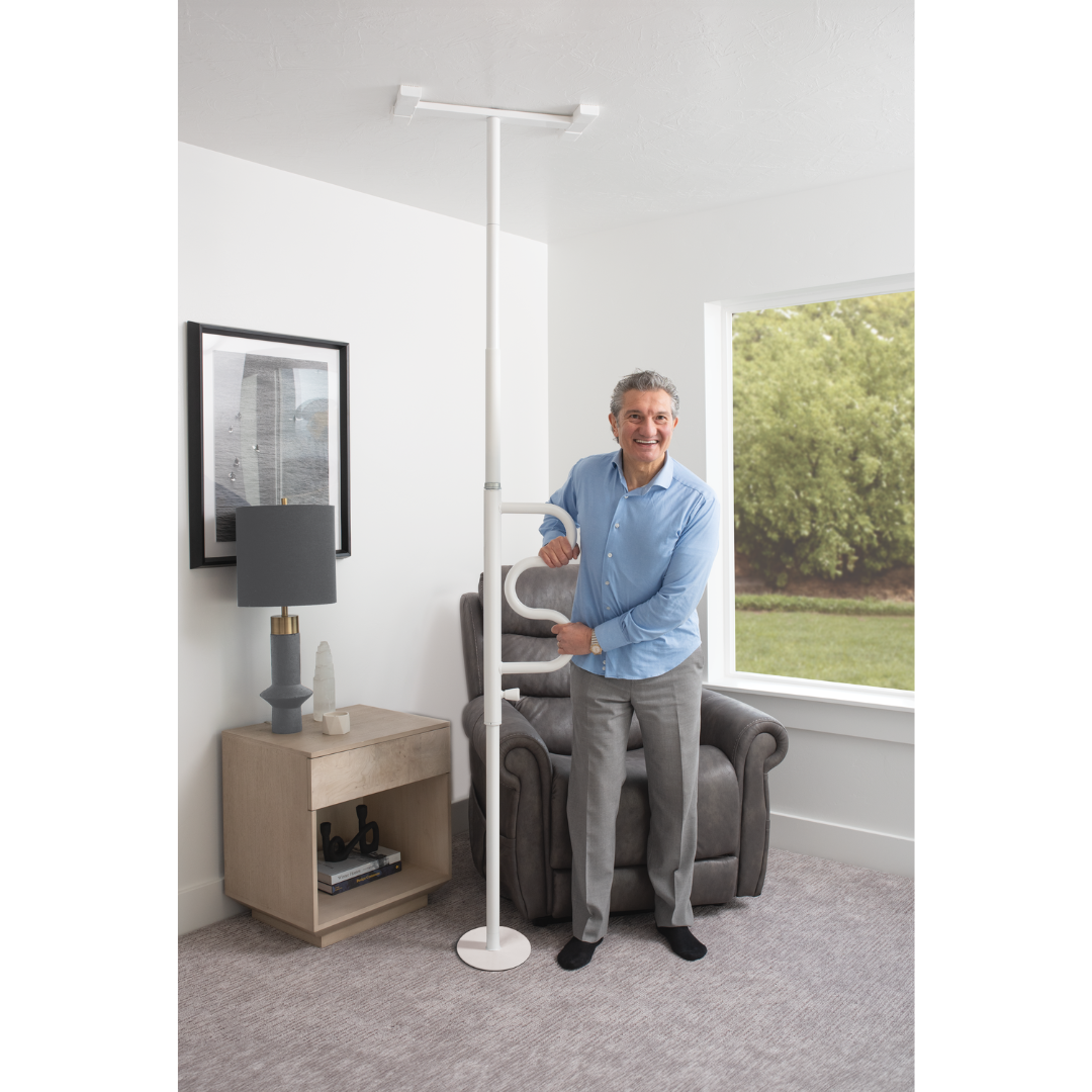 A smiling man stands by a gray armchair, holding the Stander Wonder Pole HD—Bariatric Fall Prevention Safety Pole—in a living room with a side table, lamp, and large window showing greenery outside.