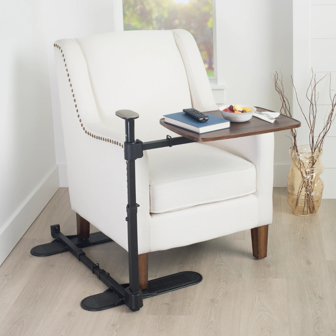 A white upholstered armchair features the Stander Wonder Tray Plus – TV Tray & Standing Aid with a wooden surface, holding a book, remote, bowl of fruit, and napkin in a bright room with light wood flooring.