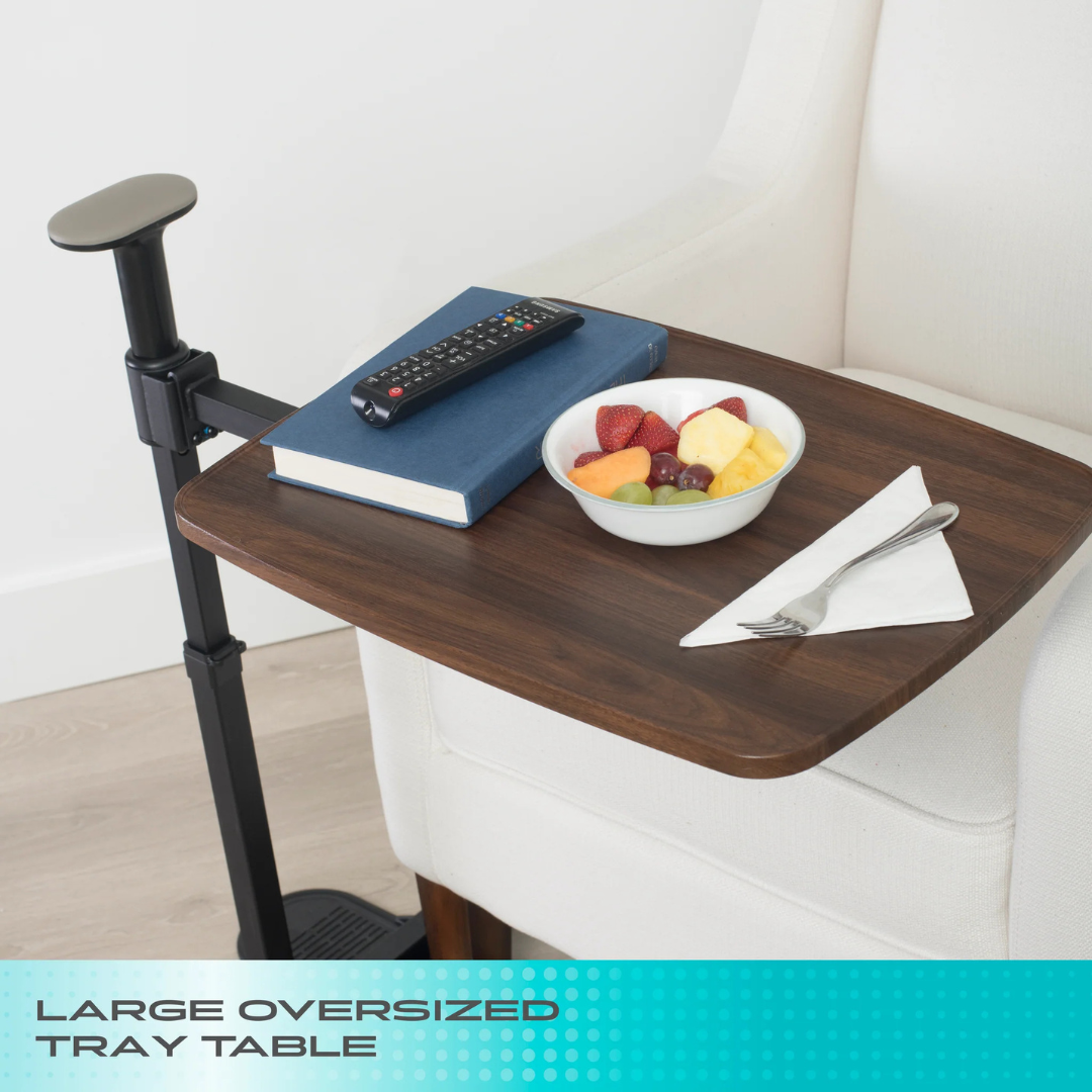 A Stander Wonder Tray Plus – TV Tray & Standing Aid, attached to a white armchair, holds a remote, a closed blue book, a bowl of mixed fruit, and a fork on a napkin. Text at the bottom reads "Large Oversized Tray Table.