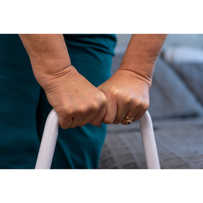 Close-up of an older adult’s hands gripping a white Step2Bed Mini Bedside Stepper rail, with a bed and grey blanket in the background. The person is wearing a gold ring and a teal outfit.