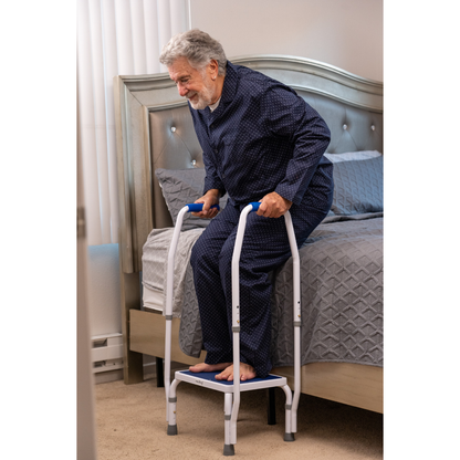 An elderly man in pajamas uses the Step2Bed Mini Bedside Stepper by his bed, gripping the handles with both feet on the platform in a softly lit bedroom, promoting safe and easy standing for fall prevention.