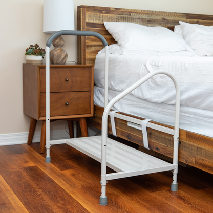The Step2Bed XL Bedside Stepper, a white metal safety step with handrails, is placed beside a wooden bed and nightstand with a lamp and plant on a hardwood bedroom floor to help prevent falls.