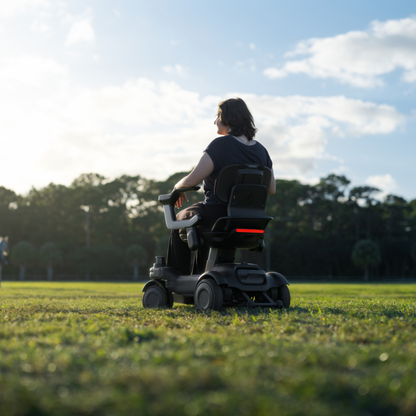 A person using a STRUTT Electric Vehicle ev1 motorized wheelchair sits on grass in a sunny, open park, facing away from the camera toward trees beneath a blue sky dotted with clouds. The STRUTT ev1 is not classified as a medical device and features guided sensors and unique innovations that go beyond standard mobility specifications.