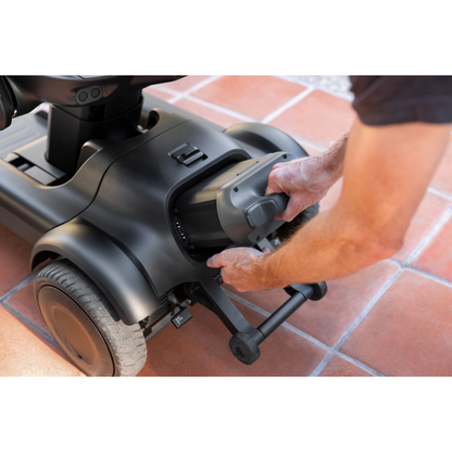 A person removes the battery from a black STRUTT Electric Vehicle ev1, which is parked on a tiled surface; this scooter, featuring advanced specifications and guided sensors for smarter driving, is intended for personal transport and is not a medical device.