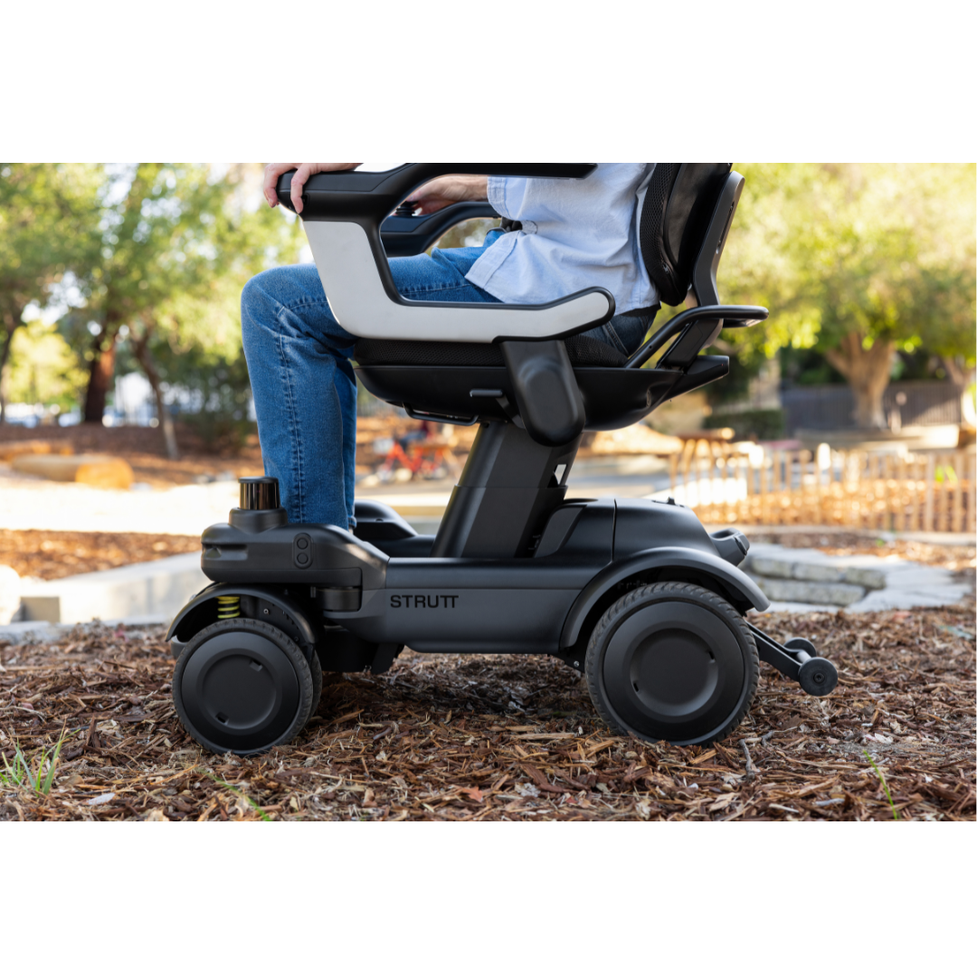 A person sits in a sleek, modern black STRUTT Electric Vehicle ev1—Drive Smarter with Guided Sensors—on a mulch path in a sunlit park surrounded by trees. Only the lower half of their body and the ev1 are visible; its innovative design clearly emphasizes enhanced mobility and style rather than serving as a conventional medical device.