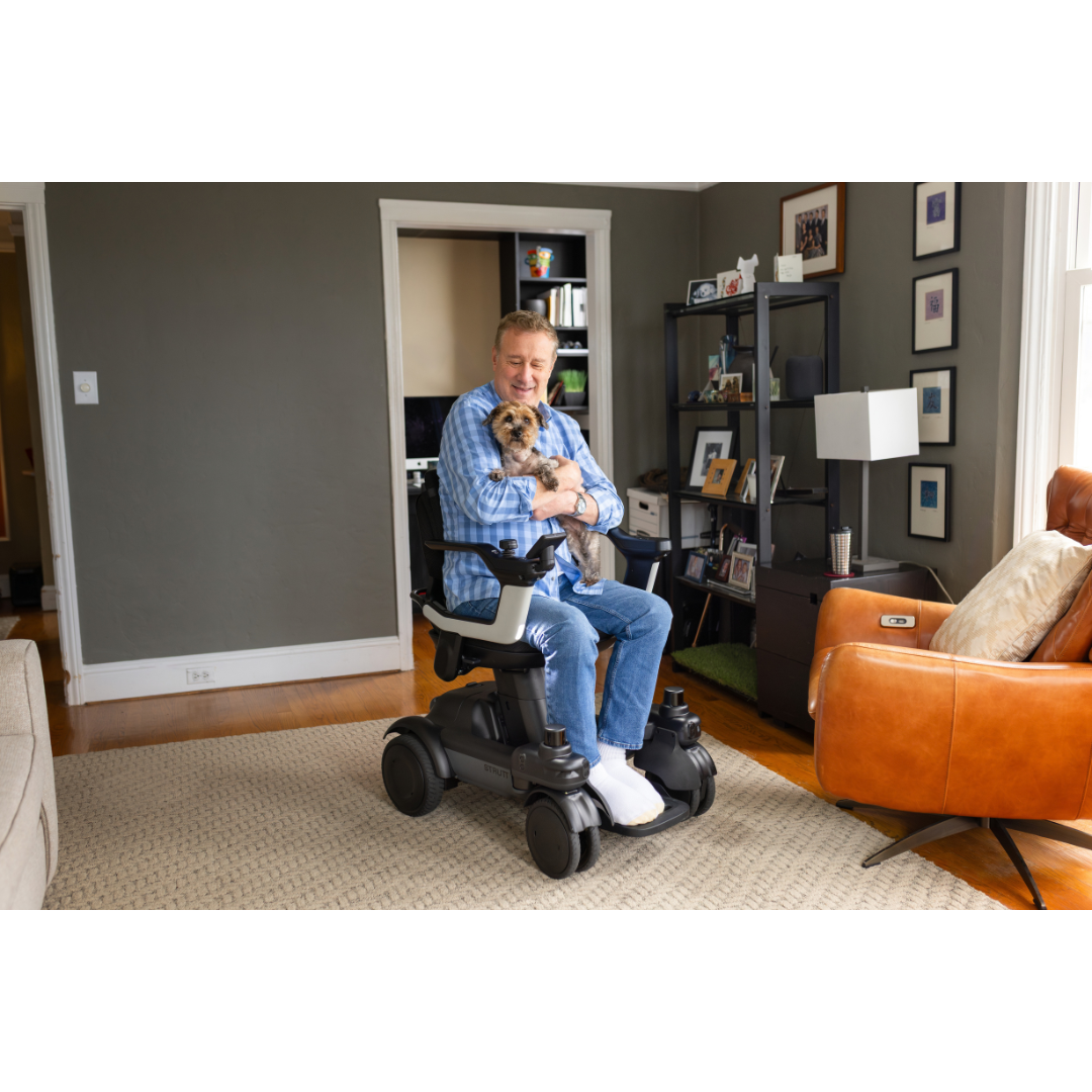 A man in the STRUTT Electric Vehicle ev1 | Power Mobility For Every Day Use holds a small dog and smiles in a cozy living room, surrounded by a brown leather chair, bookshelf, and framed photos—a perfect scene to showcase the comfort and accessibility this power mobility solution provides for everyday living.