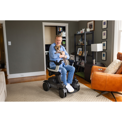 A man in the STRUTT Electric Vehicle ev1 | Power Mobility For Every Day Use holds a small dog and smiles in a cozy living room, surrounded by a brown leather chair, bookshelf, and framed photos—a perfect scene to showcase the comfort and accessibility this power mobility solution provides for everyday living.