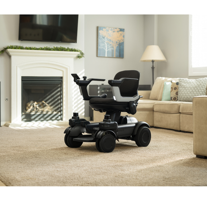 A STRUTT Electric Vehicle ev1 | Drive Smarter with Guided Sensors—showcasing a sleek black and silver design and advanced ev¹ specifications for everyday mobility (not a medical device)—sits on a beige carpet in a cozy living room with neutral decor, a fireplace, a TV, and a beige sectional sofa.