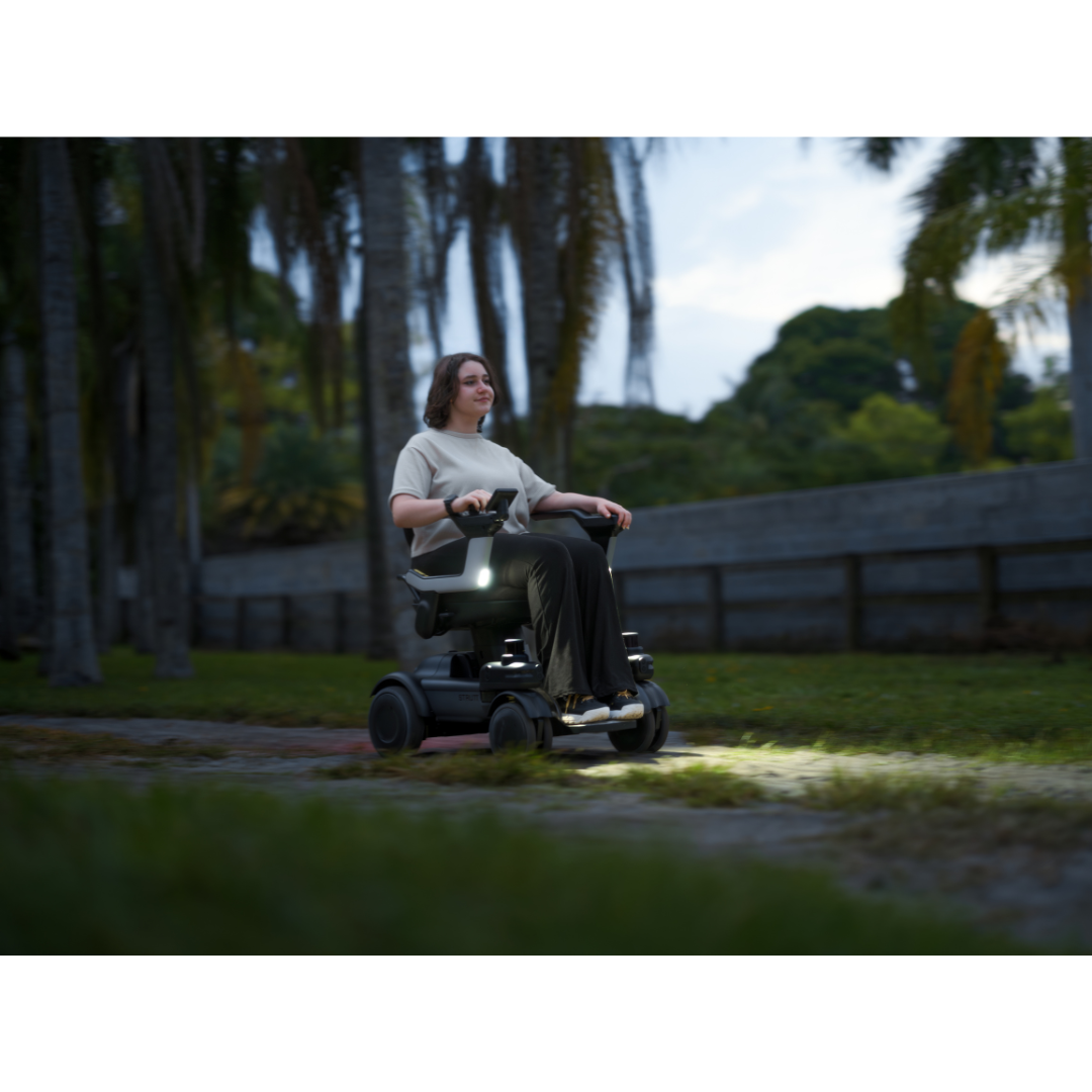 A person drives the STRUTT Electric Vehicle ev1, its front lights aglow, along an evening path lined with palm trees—demonstrating smart guided sensors and underscoring that it is not a medical device.