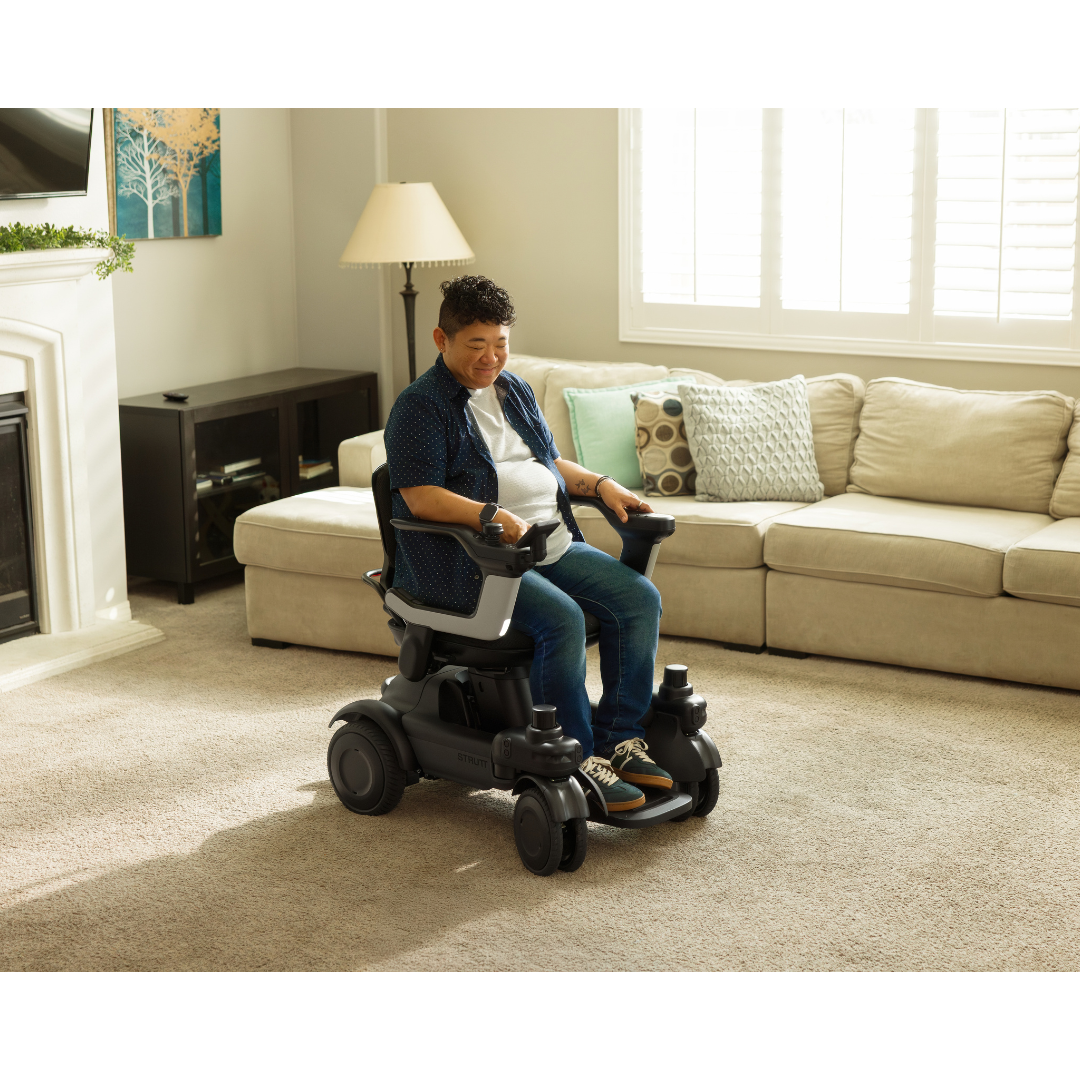 A person sits in a STRUTT Electric Vehicle ev1—an advanced model with guided sensors and modern features, distinct from traditional medical devices—in a bright living room furnished with a beige sofa, a lamp, and wall art. Sunlight streams through large windows adorned with white shutters.