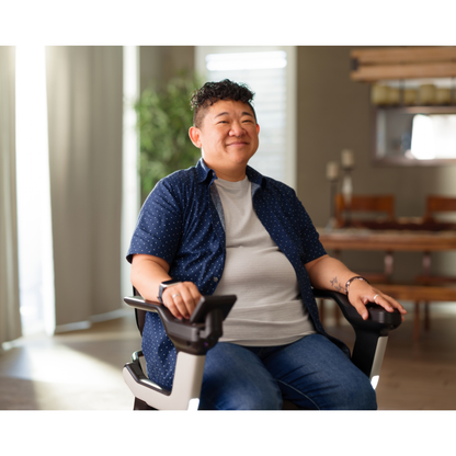 A person with short curly hair smiles while sitting indoors in a STRUTT Electric Vehicle ev1, wearing a blue shirt over a gray t-shirt and jeans. Sunlight streams through the windows behind them as they enjoy the comfort and thoughtful specifications of their chair, designed with guided sensors to help drive smarter—reminding us that this is not a medical device.