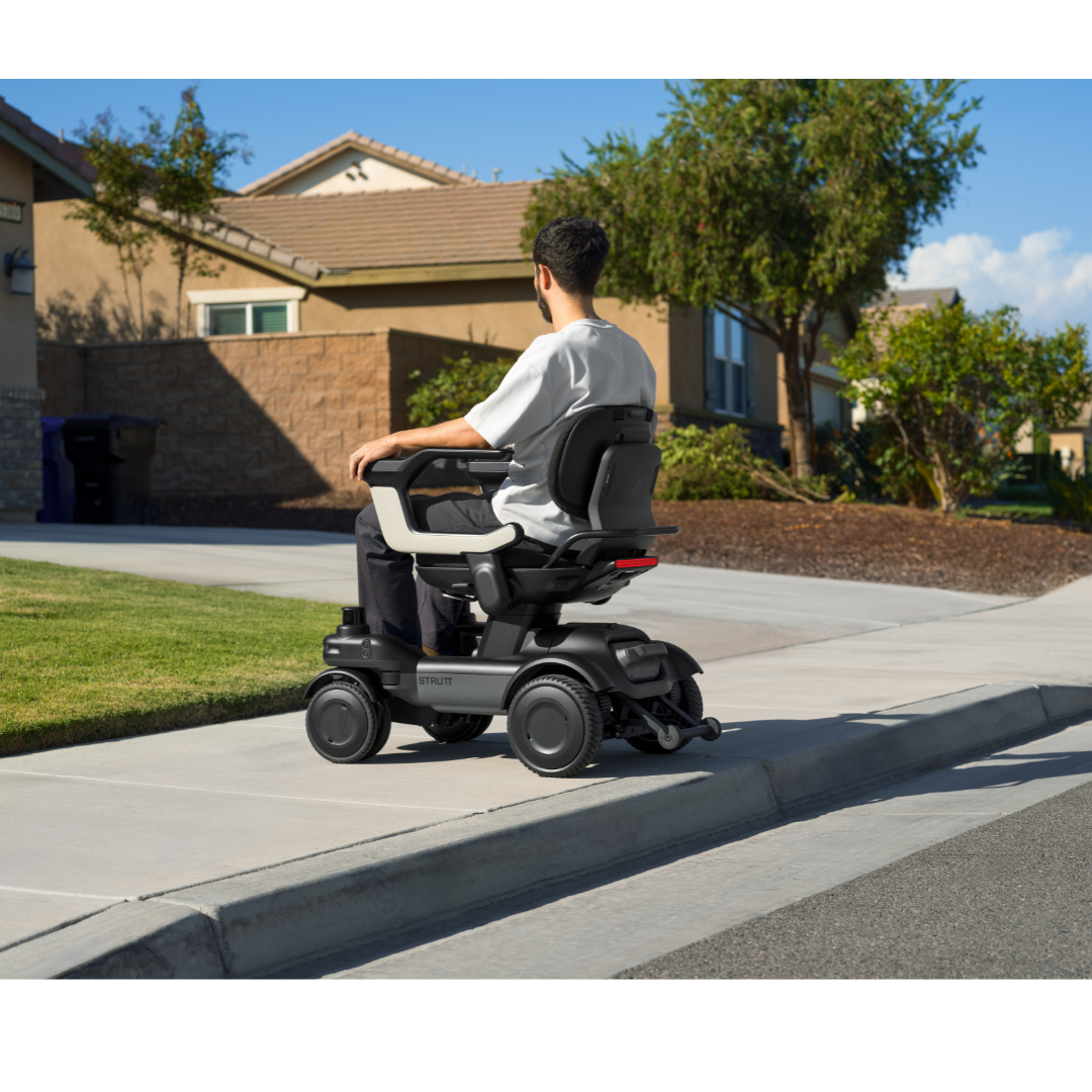 A person in a STRUTT Electric Vehicle ev1, which is not a medical device, ascends a sidewalk curb outside a suburban house on a sunny day, driving smarter with guided sensors.