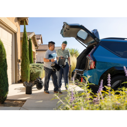 Two people stand by an SUV with its trunk open, holding equipment next to a house. On the driveway, there are containers and a STRUTT Electric Vehicle ev1 | Drive Smarter with Guided Sensors—clearly marked as not a medical device—alongside additional gear, plants, and flowers visible in the foreground. Specifications for the STRUTT ev1 appear to be under review as they prepare their setup.