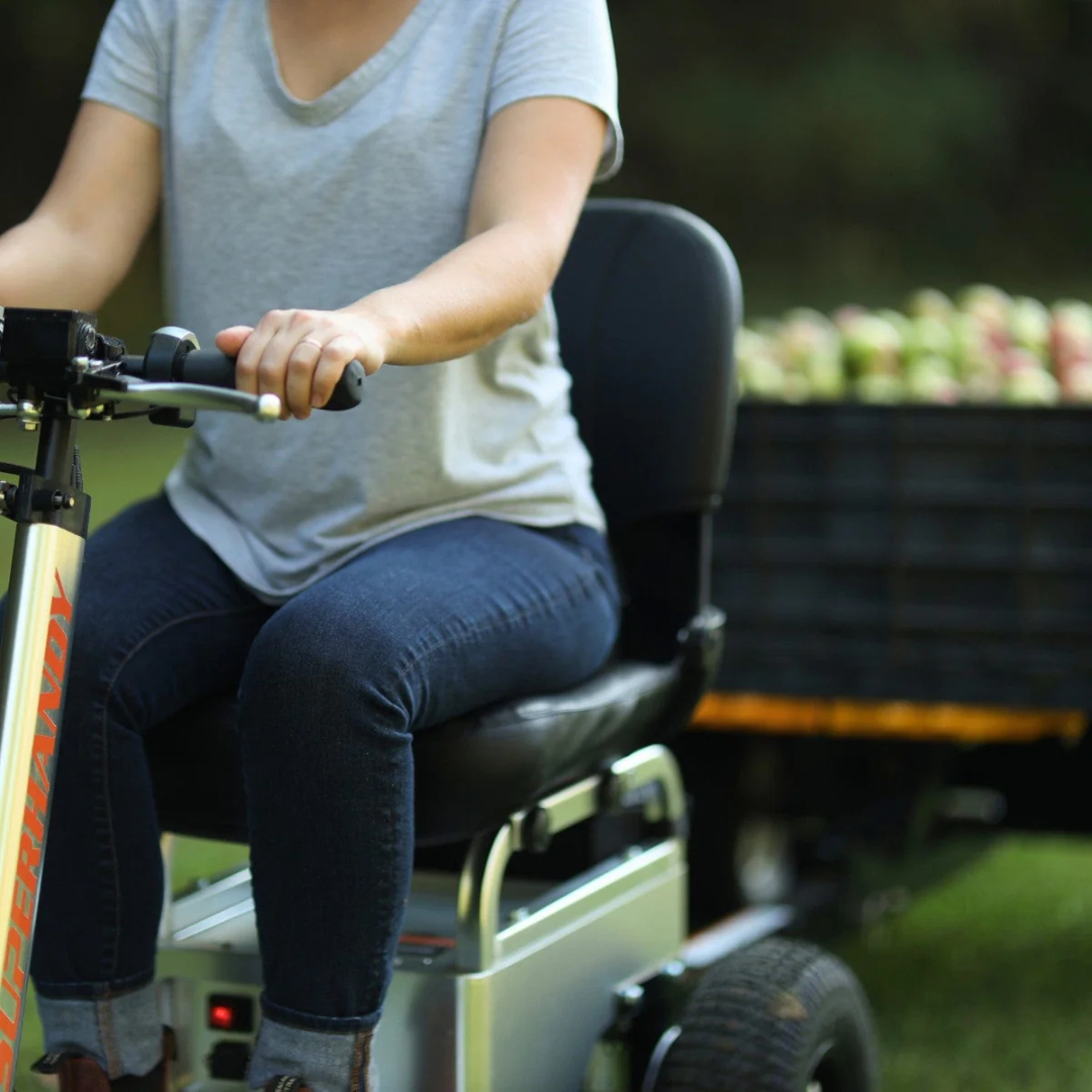 A person in a gray T-shirt and jeans sits on a SuperHandy Tugger Tow Tractor – Powerful Towing Scooter, holding the handlebars. Outdoors, a trailer filled with apples is attached to the compact tow tractor in the background.