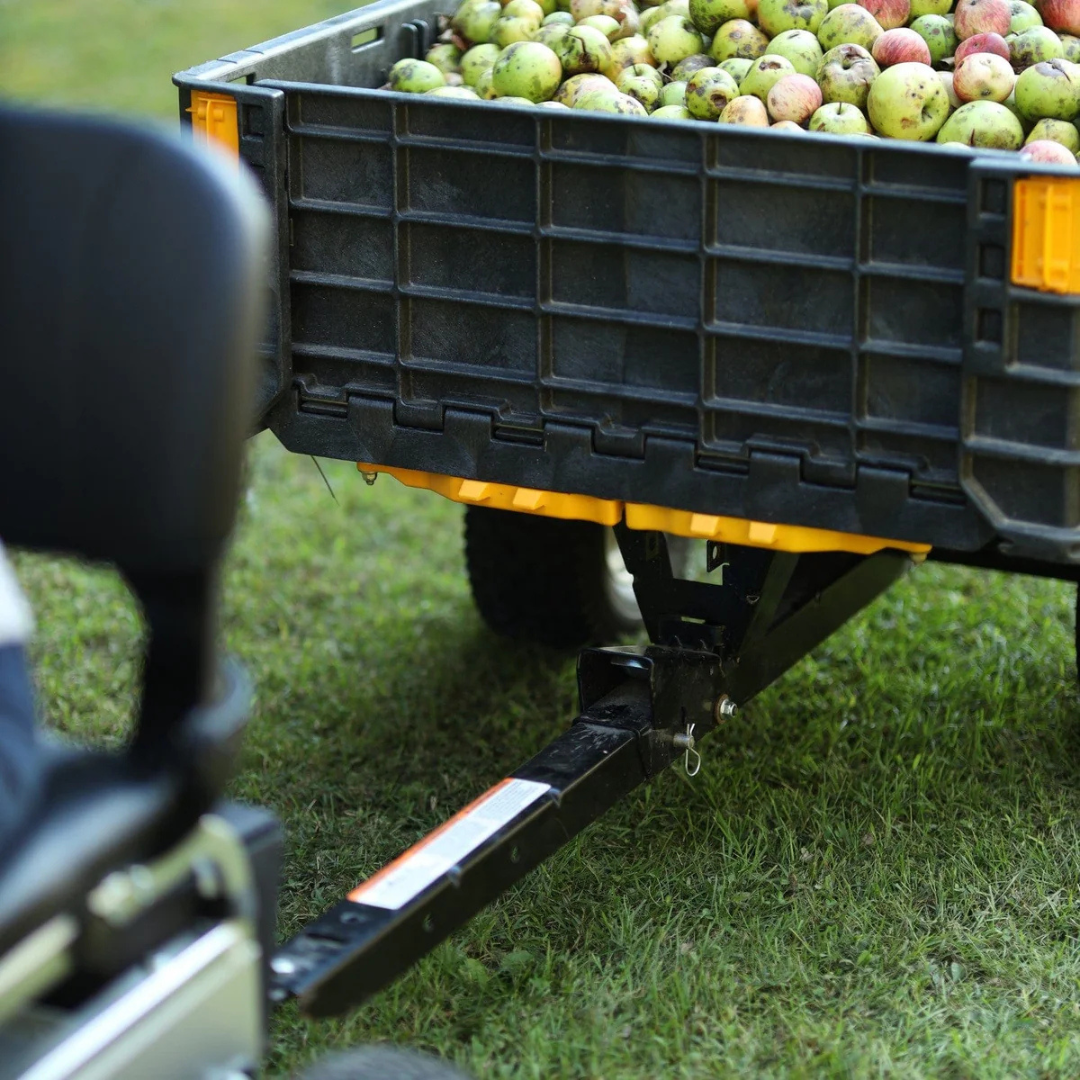 A black trailer filled with fresh apples is attached to the SuperHandy Tugger Tow Tractor - Powerful Towing Scooter, parked on green grass. The edge of a seat appears in the foreground.