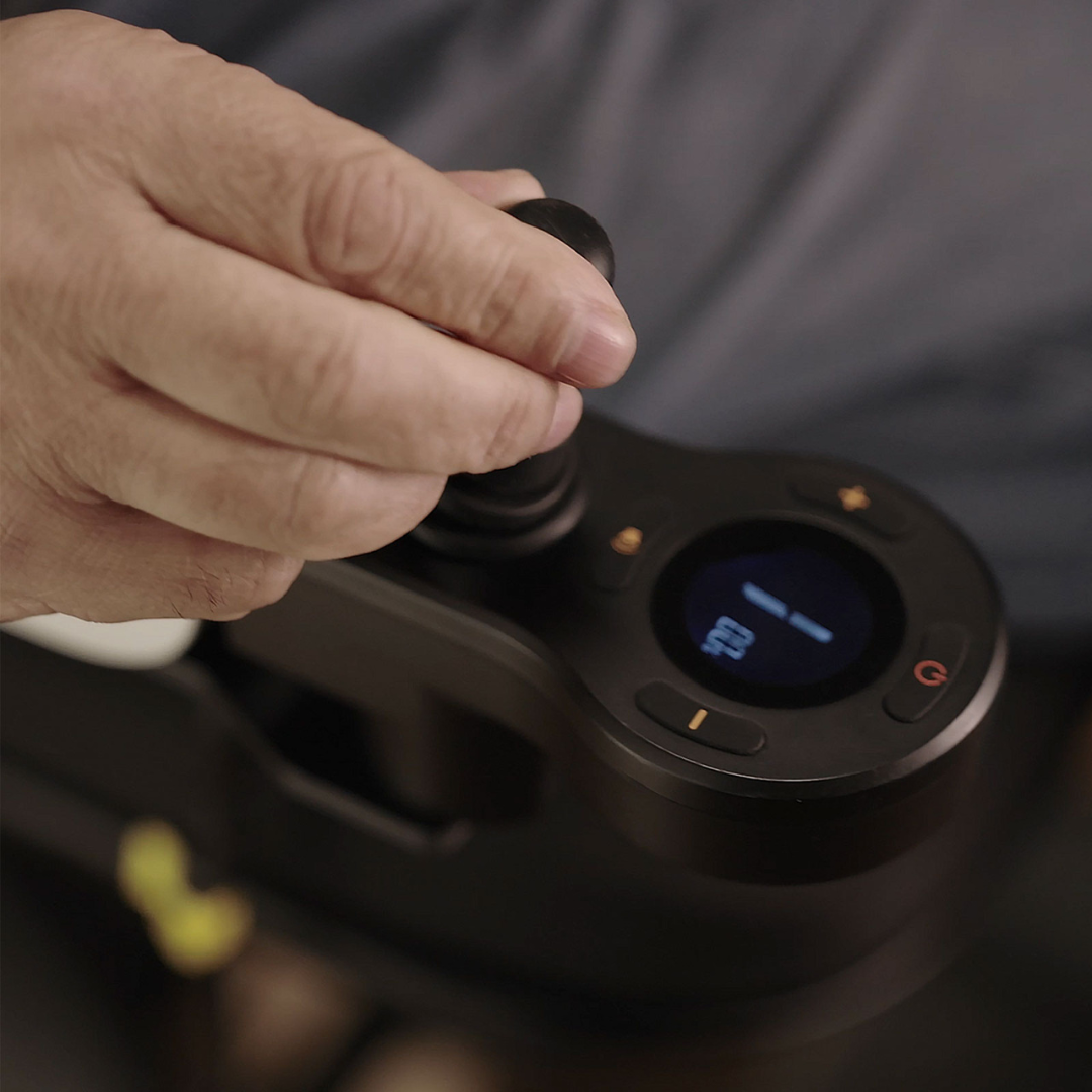 A close-up of a hand using the Superpi P2 Portable Electric Wheelchair’s black joystick with a digital display showing a wheelchair icon, highlighting essential accessibility and assistive technology features.