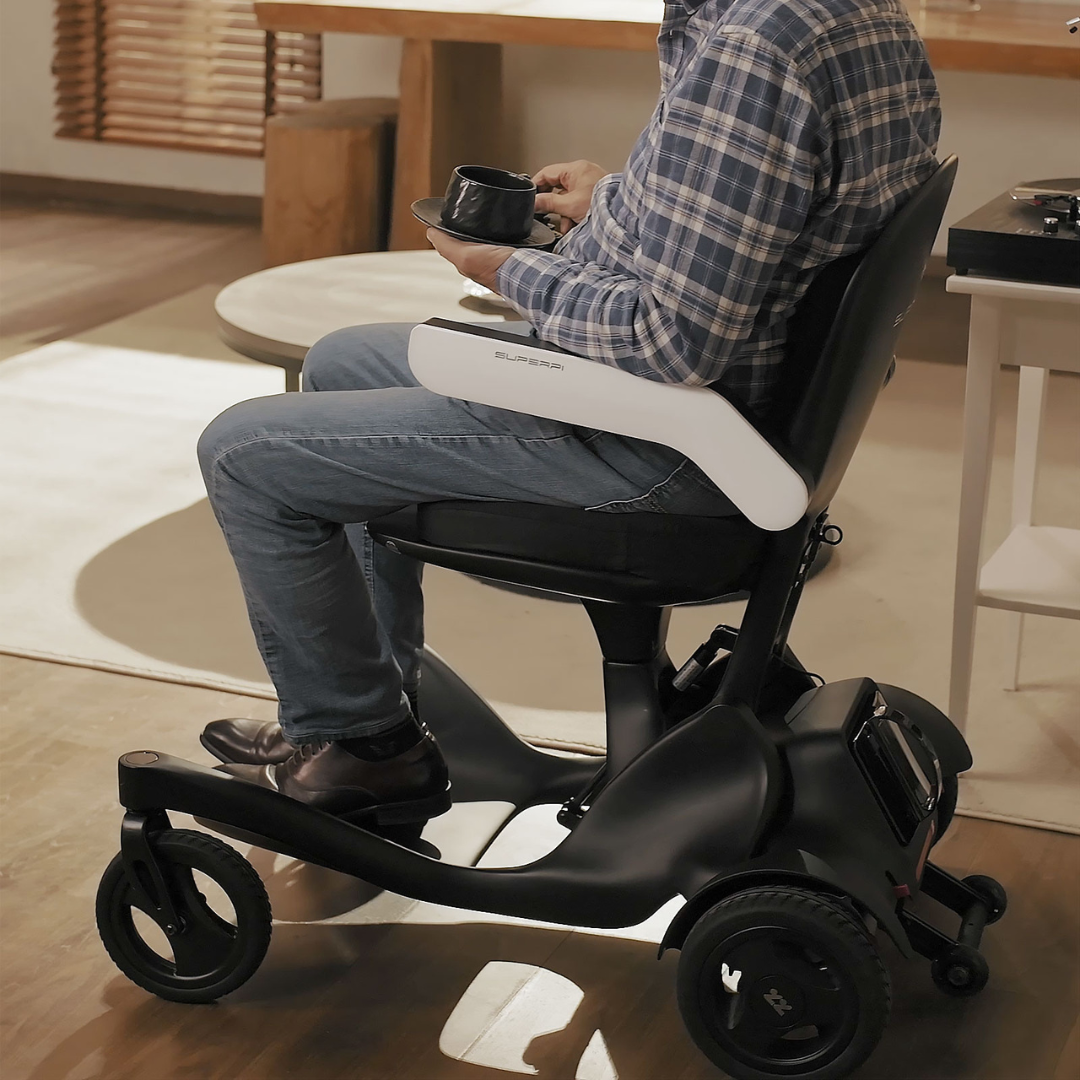 A person in a plaid shirt and jeans sits indoors in a sleek black and white Superpi P2 Portable Electric Wheelchair, holding a coffee cup. The room has warm lighting, wooden floors, and modern accessibility features.