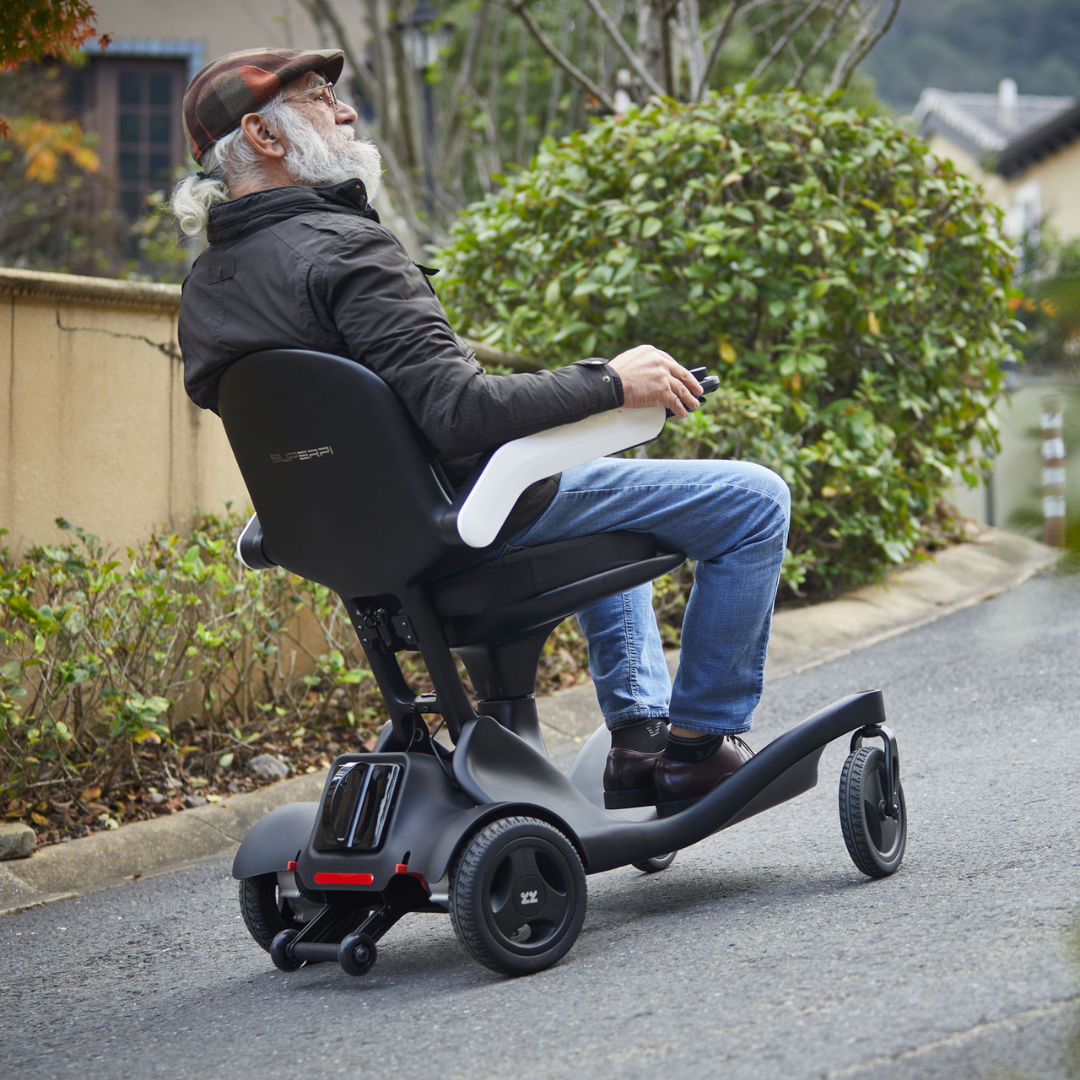 An older man with a gray beard, in a hat and jacket, rides the Superpi P2 Portable Electric Wheelchair up a sloped outdoor path lined with greenery and houses.