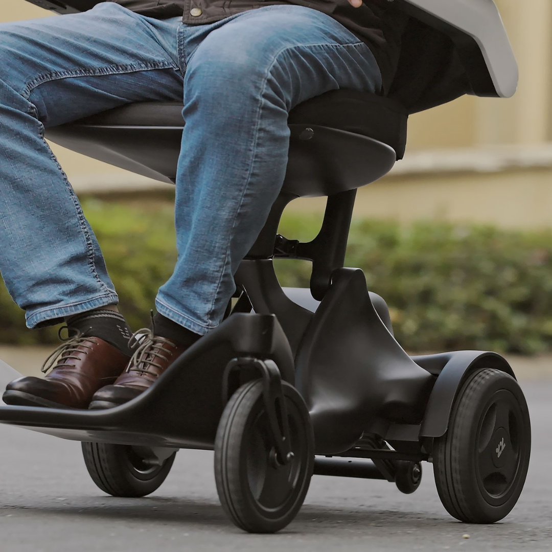 A person in blue jeans and brown shoes rides the Superpi P2 Portable Electric Wheelchair, a sleek black three-wheeler, outdoors on a paved path—enjoying smooth, comfortable, and convenient travel for daily adventures.