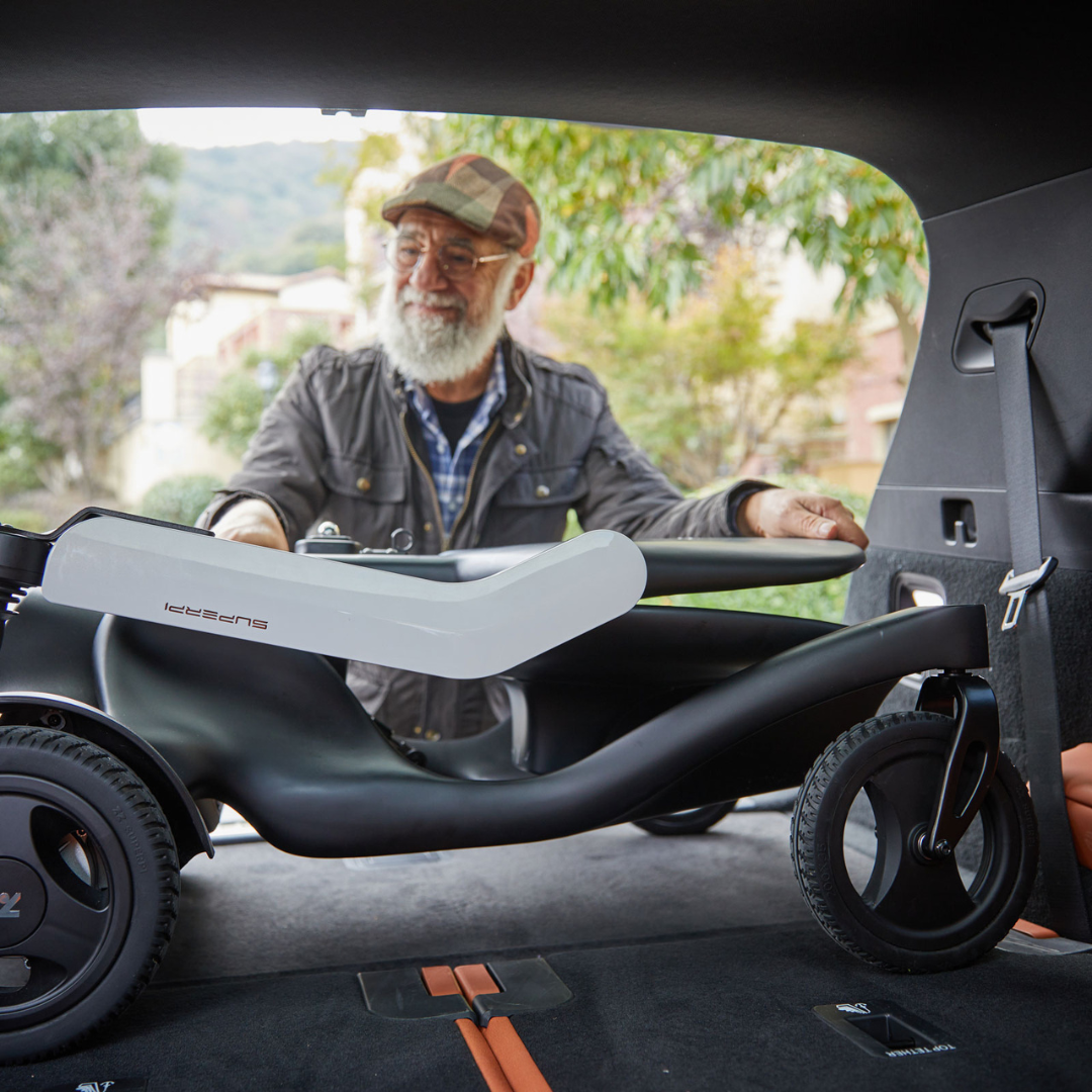 An older man with a beard and cap is loading a folded Superpi P2 Portable Electric Wheelchair into the trunk of a car, with greenery and houses visible in the background.