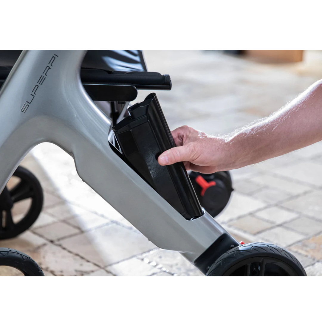 A person’s hand removing or inserting a black battery pack from the side compartment of the gray Superpi W1 Electric Wheelchair Rollator with 4-in-1 Functionality, shown on a tiled floor.