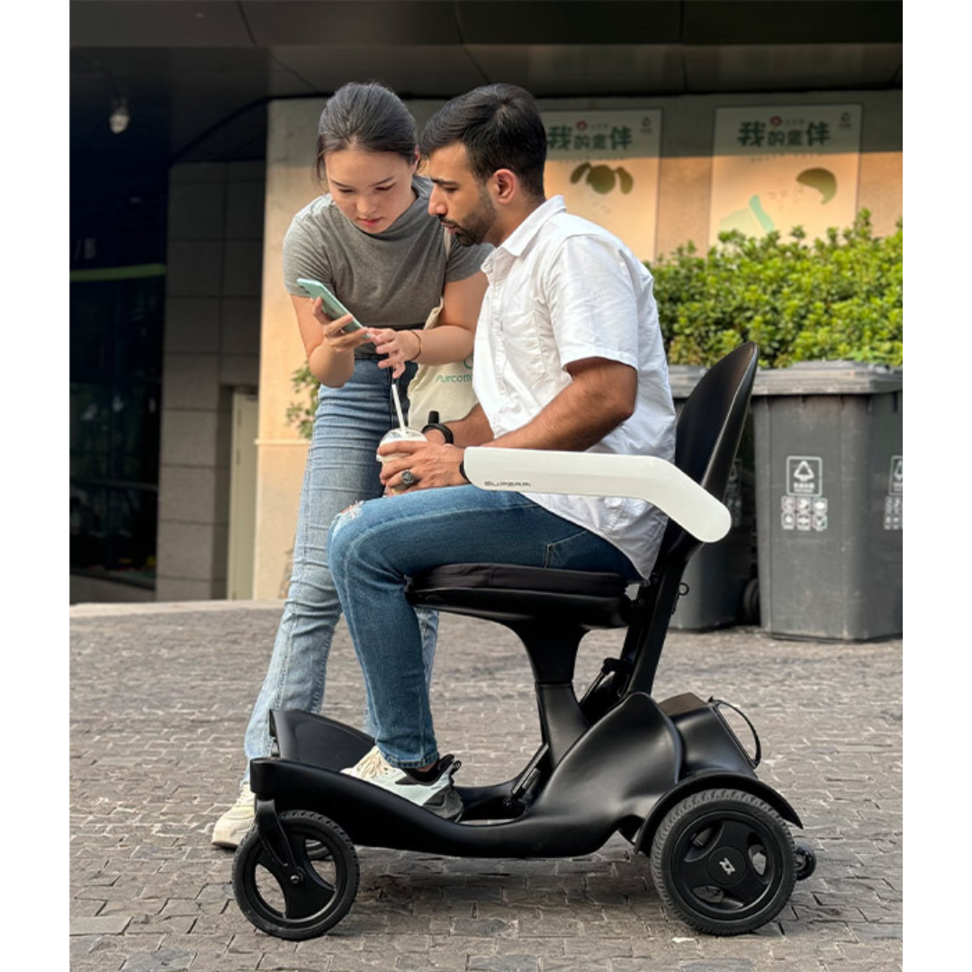 A woman shows her phone to a man outdoors. The man, dressed in a white shirt and jeans, sits on a Superpi P2 Portable Electric Wheelchair, holding a drink and listening. Buildings and trash bins can be seen in the background.