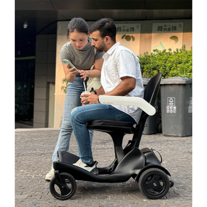 A woman shows her phone to a man outdoors. The man, dressed in a white shirt and jeans, sits on a Superpi P2 Portable Electric Wheelchair, holding a drink and listening. Buildings and trash bins can be seen in the background.