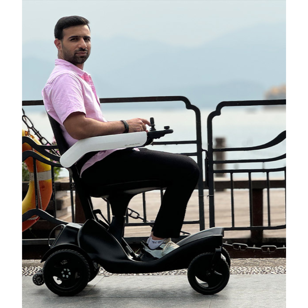A man in a pink shirt and black pants sits on a Superpi P2 Portable Electric Wheelchair near the waterfront, looking toward the camera.