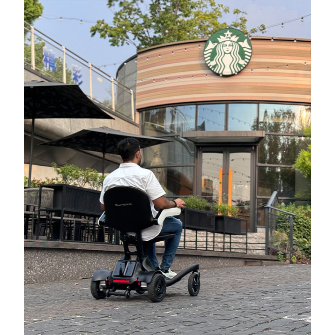 A man in a Superpi P2 Portable Electric Wheelchair faces a Starbucks café on a cobblestone path, surrounded by greenery and outdoor seating with large umbrellas. The provided product description is only CSS code, not relevant product info.