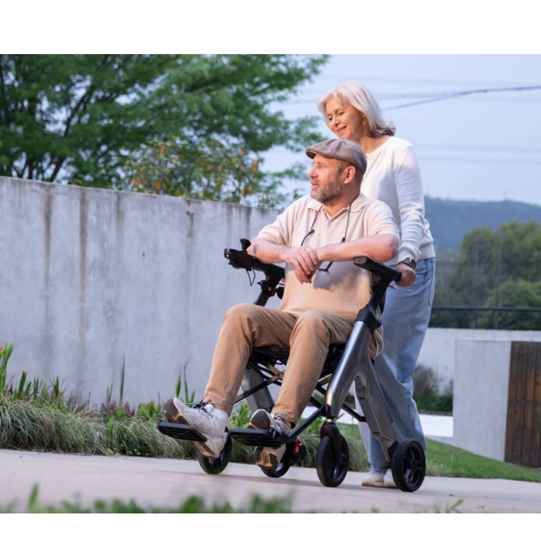 An older man uses the Superpi W1 Electric Wheelchair Rollator with 4-in-1 Functionality, while an older woman stands behind him smiling as they enjoy an outdoor walk on a paved path surrounded by greenery.