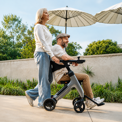 A woman pushes a man in a Superpi W1 Electric Wheelchair Rollator with 4-in-1 Functionality outdoors on a sunny day. Both look happy, with green trees and large white umbrellas in the background.