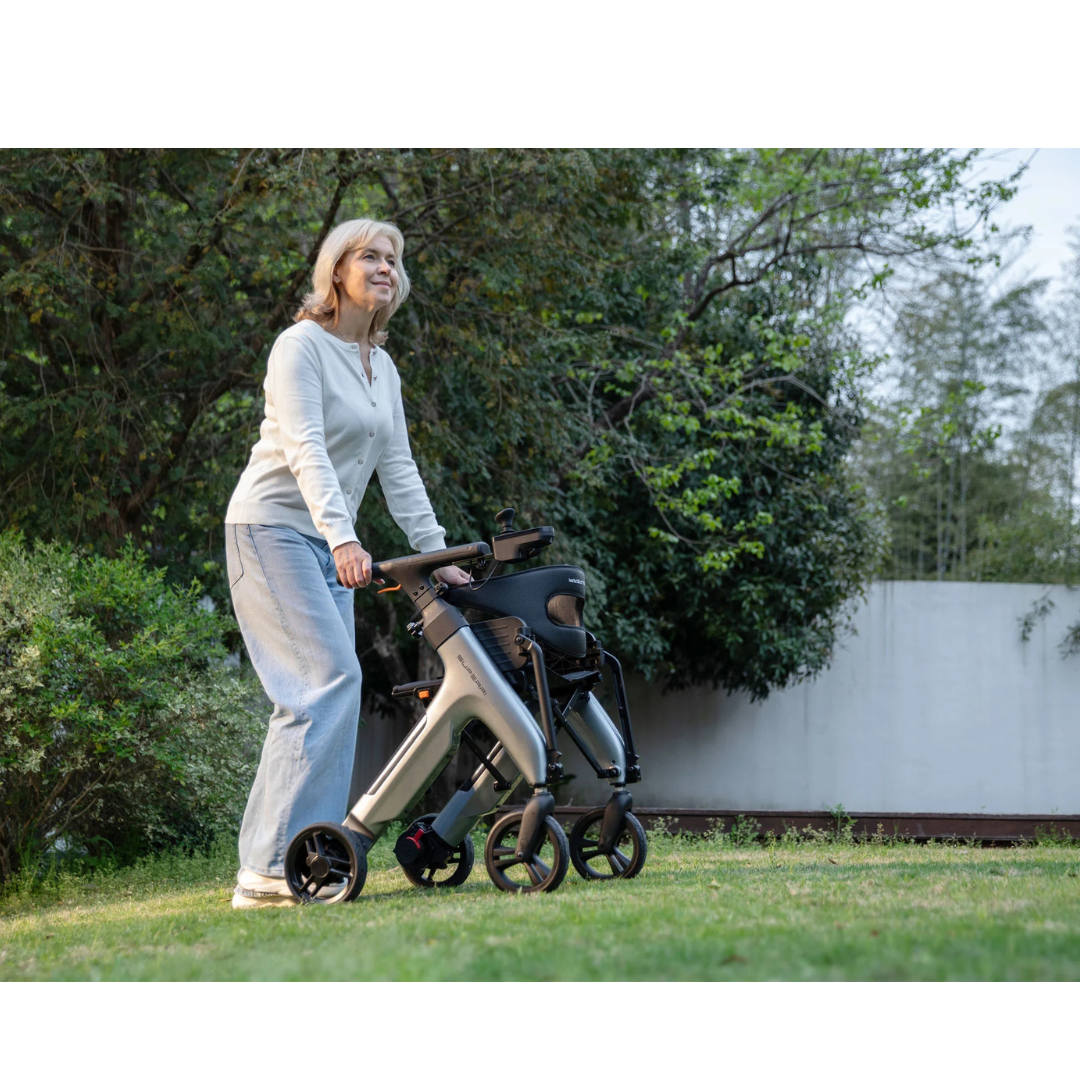 An older woman with gray hair smiles while walking outdoors on grass using the Superpi W1 Electric Wheelchair Rollator with 4-in-1 Functionality. She wears a light sweater and jeans, surrounded by green trees and bushes.