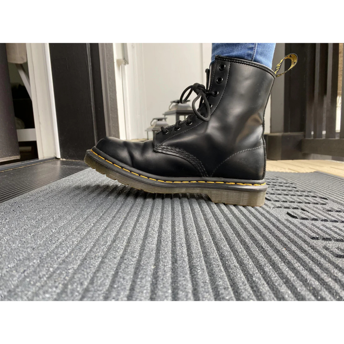 A person in black leather lace-up boots with thick soles and yellow stitching stands on an EZ-Access TRANSITIONS® Angled Entry Welcome Mat by a doorway, seamlessly transitioning into the room.