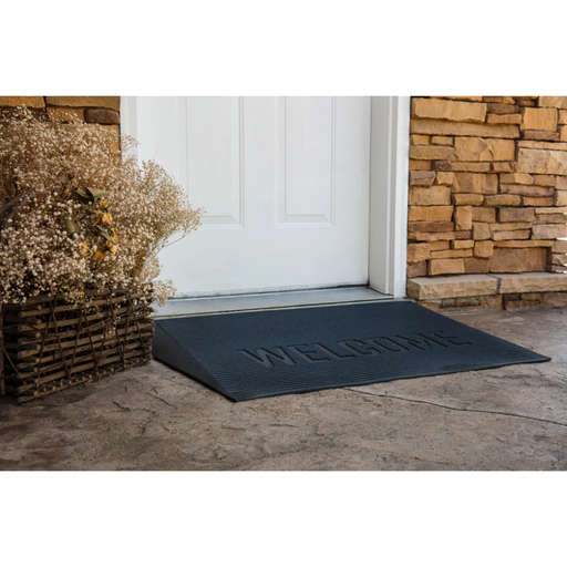 The EZ-Access TRANSITIONS® Angled Entry Welcome Mat, featuring Welcome, is at the white door entrance. A wicker basket filled with dried flowers sits to the left, and its slip-resistant surface offers safety as you step onto the concrete floor of the stone-walled entrance.