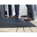 Wearing brown shoes and dark pants, a person steps onto an EZ-Access TRANSITIONS® Angled Entry Welcome Mat with a slip-resistant surface, providing stability on the black textured mat placed before the doorway on a wooden floor.