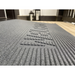 Close-up of an EZ-Access TRANSITIONS® Angled Entry Welcome Mat in gray with a textured, slip-resistant surface and raised patterns on a wooden floor. In the background, three lanterns rest against the wall.