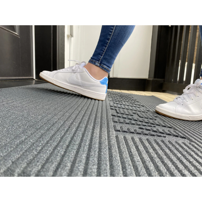 A person in white sneakers and blue jeans steps onto an EZ-Access TRANSITIONS® Angled Entry Welcome Mat near a dark door. The focus is on the shoes and the slip-resistant mat, with part of the door and floor visible in the background.