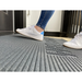 A person in white sneakers and blue jeans steps onto an EZ-Access TRANSITIONS® Angled Entry Welcome Mat near a dark door. The focus is on the shoes and the slip-resistant mat, with part of the door and floor visible in the background.