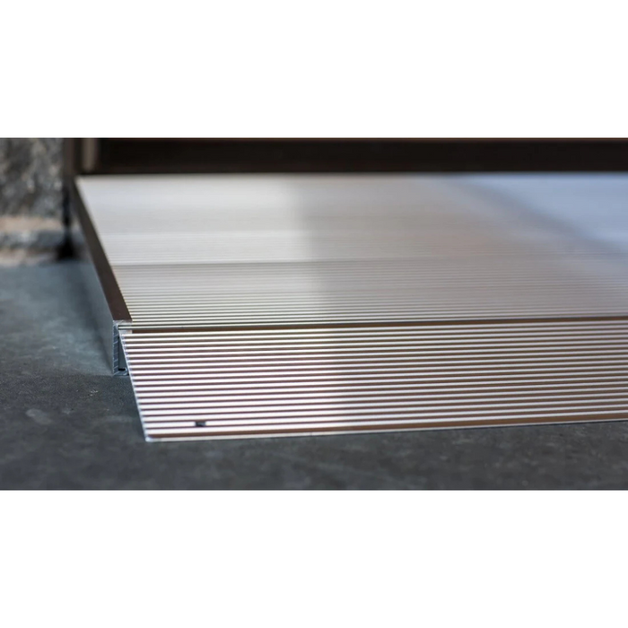 The EZ-Access TRANSITIONS® Angled Entry Ramp with Adjustable Legs features a ribbed surface for traction and is installed over doorways to ensure smooth transitions between floor levels. Its aluminum design optimizes placement against a stone background.