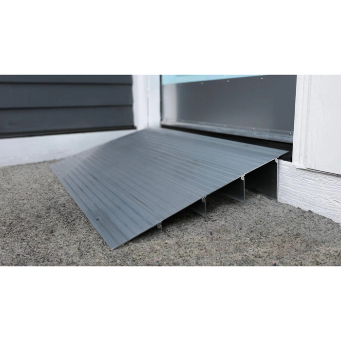 An EZ-Access TRANSITIONS® Modular Entry Ramp, made of silver metal and slip-resistant, is placed at a doorway. It connects the buildings exterior stone surface to its interior, with the dark gray siding contrasting with the white door frame for seamless wheelchair accessibility.