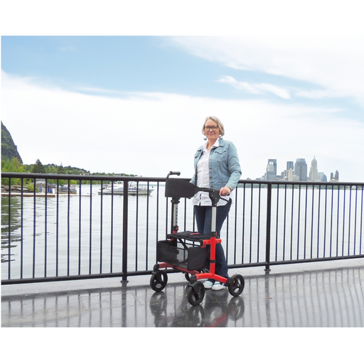 A person with short blonde hair stands on a boardwalk near a railing, holding a Triumph Mobility Escape Rollator - One Handle Fold, weighing only 15 lbs. The backdrop features water and a city skyline under a blue sky.