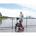 A person with short blonde hair stands on a boardwalk near a railing, holding a Triumph Mobility Escape Rollator - One Handle Fold, weighing only 15 lbs. The backdrop features water and a city skyline under a blue sky.