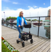 A woman walks beside the marina on a wooden boardwalk with her Triumph Mobility Escape Rollator, featuring an advanced braking system. She wears a blue jacket and jeans under clear skies as boats bob behind her and vibrant flowers line her path.