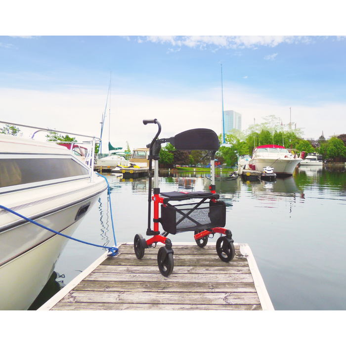 A Triumph Mobility Escape Rollator in red and black sits on a wooden dock by moored boats at a marina, set against peaceful water and sailboats under a clear blue sky, showcasing its compact one-handle folding system for convenient storage.