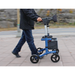 A person clad in dark attire easily maneuvers a Triumph Mobility Escape Rollator on the sidewalk. The blue and black design contrasts with the blurred street and rocky surface, highlighting its smooth glide aided by the advanced braking system and compact 15-lb foldable frame.