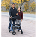 A man confidently uses his Triumph Mobility Escape Rollator - One Handle Fold, weighing just 15 lbs, as he and a woman stroll along a tree-lined path. Both smile in the autumn setting, with leaves crunching underfoot against a rocky backdrop.