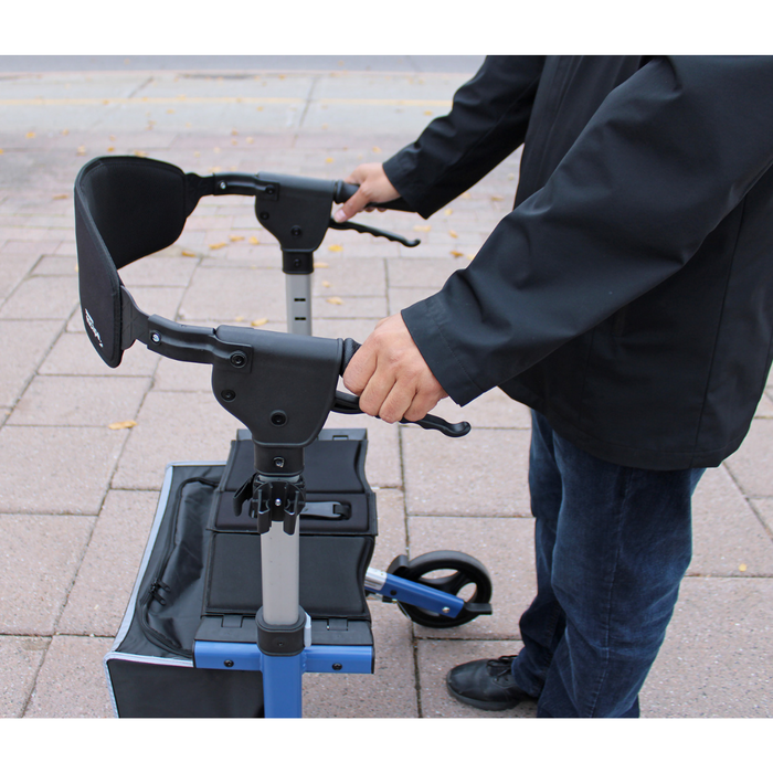 A person in a black jacket holds the handles of a blue Triumph Mobility Escape Rollator on a paved sidewalk. This 15 lb walker features one-handle fold, an advanced braking system, padded seat, and storage compartment. Fallen leaves are scattered on the ground.